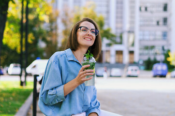Fototapeta premium Urban portrait of young elegant business woman in eyeglasses and casual clothes sitting on a bench in the city. Drinking healthy smoothie, reading a book and working.