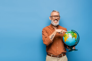 happy middle aged teacher in glasses pointing with finger at globe on blue