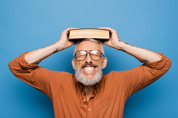 joyful middle aged teacher in glasses holding book on head on blue