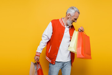 cheerful middle aged man in bomber jacket holding shopping bags on yellow