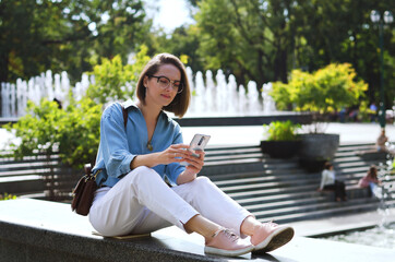 Urban portrait of young attractive business woman in eye glasses and casual clothes. Woman walking in the city street, park, talking on her phone, making selfie, works in sunny day.