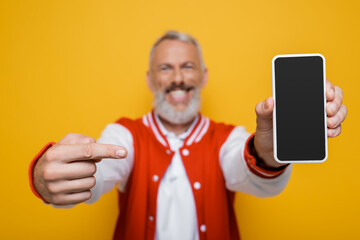 blurred and happy middle aged man pointing at smartphone with blank screen isolated on yellow