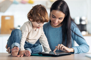 Fototapeta premium Young smiling mother and her cute son watching cartoons on digital tablet in the kitchen at home.
