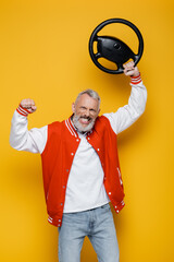 happy middle aged man in bomber jacket holding steering wheel above head on yellow