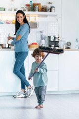 Little cute boy sweeping and cleaning the floor while his mother cooking in the kitchen at home.