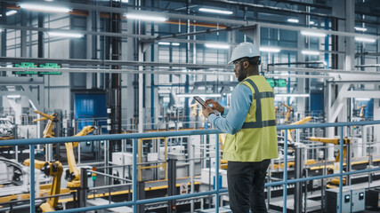 African American Car Factory Engineer in High Visibility Vest Using Tablet Computer. Automotive Industrial Manufacturing Facility Working on Vehicle Production on Automated Technology Assembly Plant.