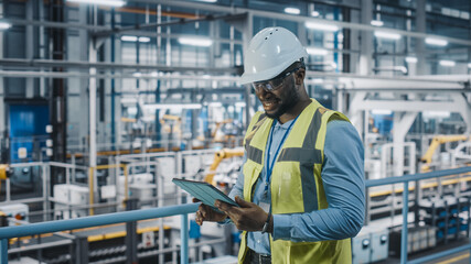 African American Car Factory Engineer in High Visibility Vest Using Tablet Computer. Automotive Industrial Manufacturing Facility Working on Vehicle Production on Automated Technology Assembly Plant.