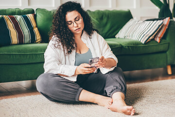 Curly haired plus size young woman wearing comfortable clothes in glasses clicks on smartphone screen sitting on floor mat in stylish room - obrazy, fototapety, plakaty