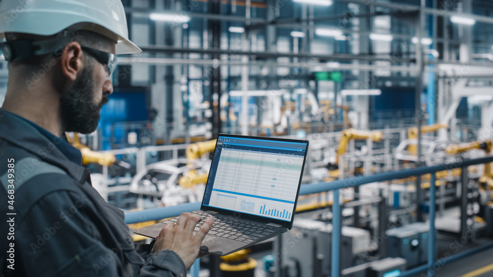 Fototapete Car Factory Engineer in Work Uniform Using Laptop Computer with Spreadsheet Software. Working with Software at Automotive Industrial Manufacturing Facility Dedicated for Vehicle Production.