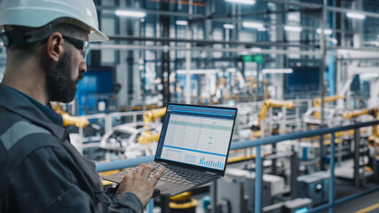 Car Factory Engineer in Work Uniform Using Laptop Computer with Spreadsheet Software. Working with Software at Automotive Industrial Manufacturing Facility Dedicated for Vehicle Production. - obrazy, fototapety, plakaty