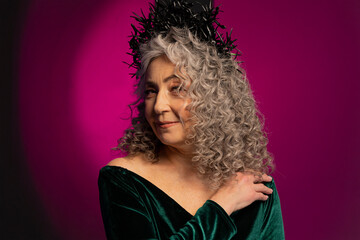 An elderly gray-haired elegant woman of 60-65 years old poses on a colored background, long curly hair. A stylish mature woman in a green dress with a deep neckline looks into the camera and smiles.