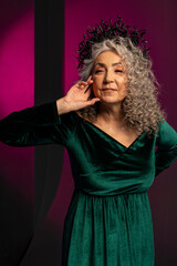 An elderly gray-haired elegant woman of 60-65 years old poses on a colored background, long curly hair. A stylish mature woman in a green dress with a deep neckline looks into the camera and smiles.