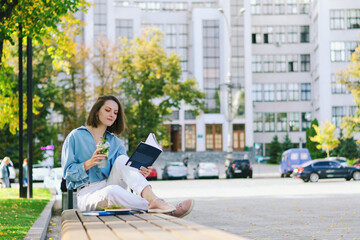 Urban portrait of young elegant business woman in eyeglasses and casual clothes sitting on a bench in the city. Drinking healthy smoothie, reading a book and working.