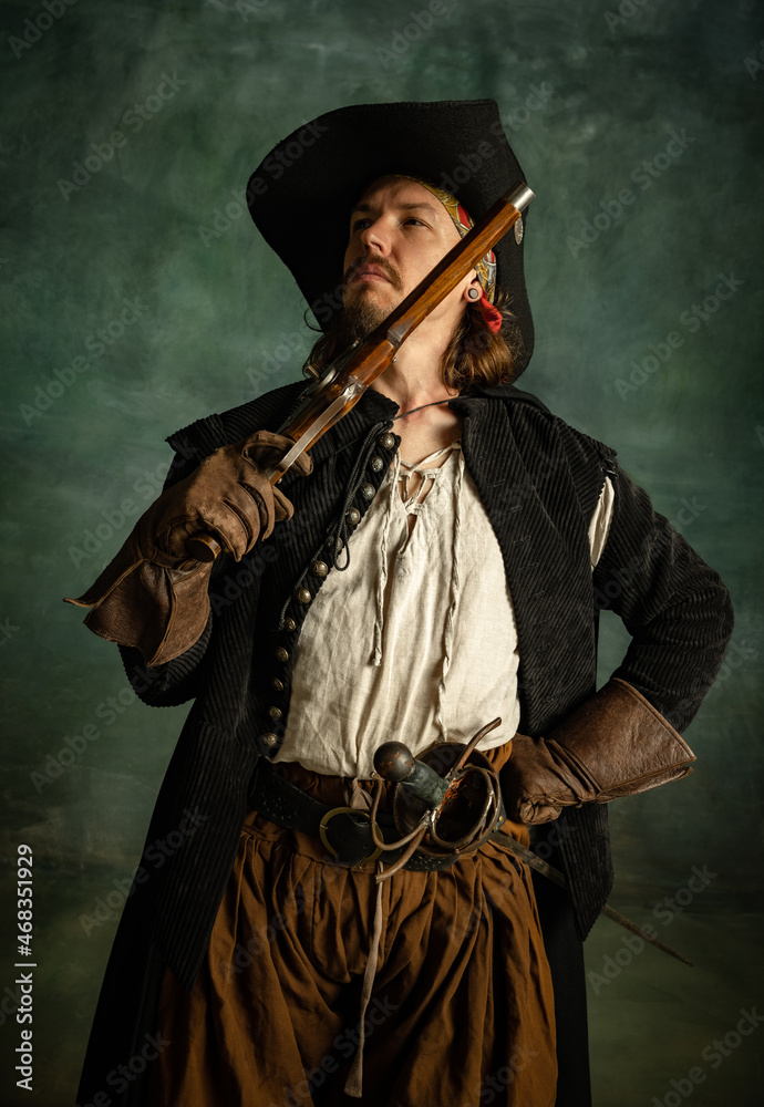 Wall mural cropped portrait of brutal man, medeival pirate in vintage costume with pistol, gun isolated over da