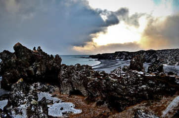 black basalt rocks covered with snow on the beach in the background sunbeams among the clouds Lóndrangar, Hellnar, Iceland