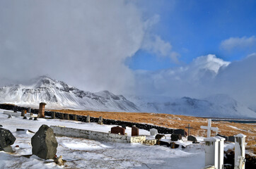 cemetery in Budir stone fence and tombstones in the background snow-capped and misty mountains, Iceland