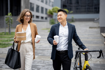 Asian businessman and caucasian businesswoman walking, drinking coffee and talking in city. Concept of business cooperation. Idea of freelance and remote work. Man with bicycle. Young woman with food