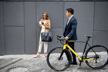 Fototapeta na wymiar Asian businessman and caucasian businesswoman on city street. Asian guy passing by with a bicycle, woman using phone. Concept of business people during a break outdoors. Modern successful people