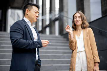 Asian businessman and smiling caucasian businesswoman smoking cigarettes during break at work on city street. Concept of modern successful business people. Idea of rest and break on job