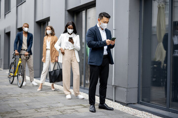 Multiracial businesspeople in medical masks waiting in queue on city street. Concept of business during Coronavirus pandemic. Idea of break on job. Businessmen and businesswomen wearing formal wear