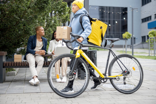 Courier walking with bicycle on background of working business people in city. Concept of shipping and logistics. Remote and freelance business work. Businessman and businesswoman talk and use laptop