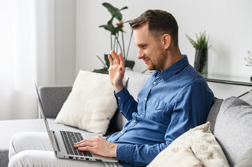 A smiling young man is sitting on the comfy couch in the living room, relaxing and talking to his friend or family, waving at the laptop screen, staying connected during pandemic