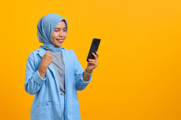 Cheerful young Asian woman looking at mobile phone isolated over yellow background