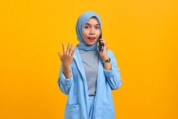 Surprised young Asian woman talking on mobile phone and raised hands isolated over yellow background