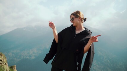 A happy young girl in sunglasses and a black hoodie stands against the background of mountains and dances