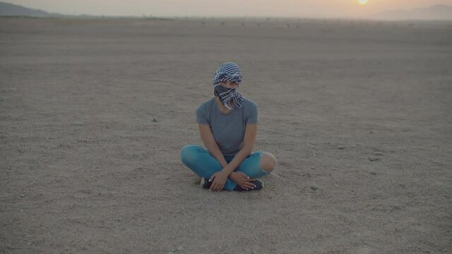 Woman in checkered keffiyeh sitting in the desert looking at camera. Caucasian female tourist sitting on desert sand.