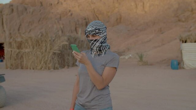 Female tourist in checkered keffiyeh standing in the desert using mobile phone.