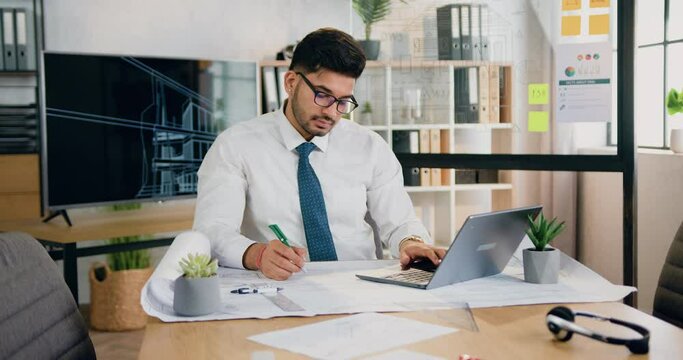 Office work concept where attractive concentrated responsible skilled bearded worker in glasses writing notes on blueprint comparing datas with information on laptop