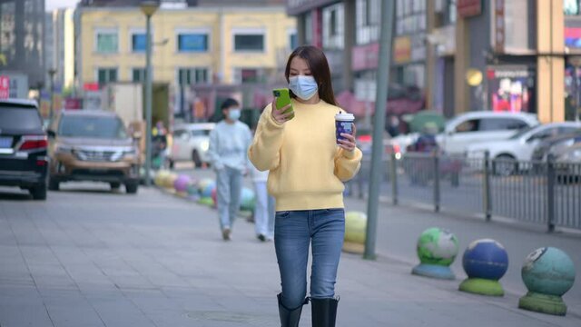 Beautiful Chinese Asian young woman walks on street during sunny autumn day. Holds drinks cup of coffee, takes pictures selfie using mobile cell phone. Wearing face mask to prevent respiratory disease