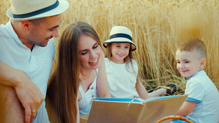 Happy family relaxing at picnic in wheat field in summer. Parents and children sitting on blanket,...