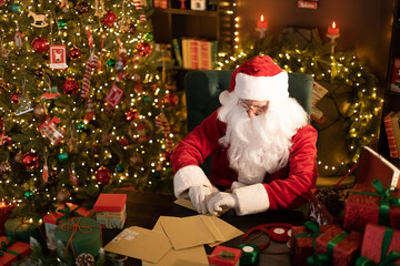 Fototapeta na wymiar Santa Clause is prepares gifts for children for Xmas at his desk while opens letters with wishes 