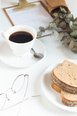 Cup of coffee, bread, menu, eucalyptus, glasses on white table. 