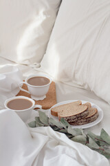 Breakfast on dark wooden vintage tray in bed with light beige sheet and pillows. Flat lay, top view. Slow romantic morning concept.