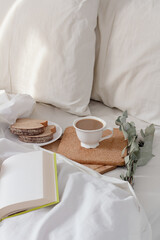 Breakfast on dark wooden vintage tray in bed with light beige sheet and pillows. Flat lay, top view. Slow romantic morning concept.