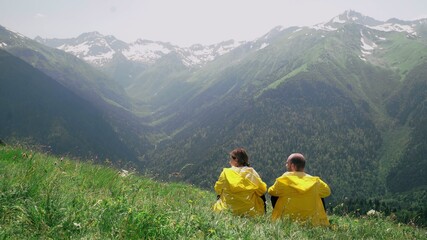 A young man and a woman in yellow raincoats are lying on the slope, enjoying the scenery of the mountainous area. Travel and tourism