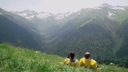 A young man and a woman in yellow raincoats are lying on the slope, enjoying the scenery of the mountainous area. Travel and tourism