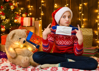 Child girl as Santa helper posing in new year decoration. Wearing a protective face mask against...