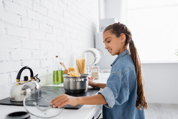 Side view of positive kid taking cap near saucepan with macaroni on stove in kitchen