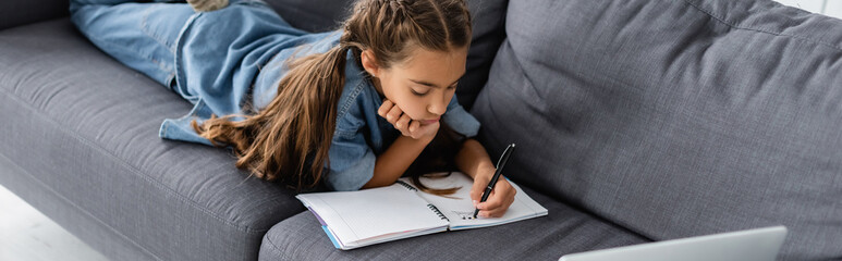 Schoolgirl writing on notebook near laptop on couch at home, banner