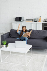 Preteen kid with notebook raising hand near laptop on couch at home