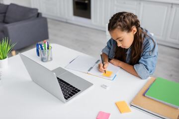 Preteen kid writing on sticky note on notebook near laptop at home