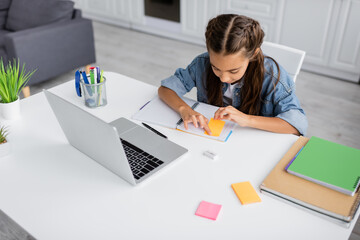Girl applying sticky note on notebook near stationery and laptop at home