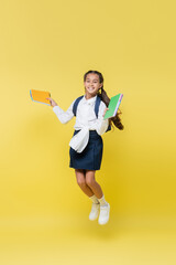 Smiling schoolgirl with notebooks jumping and looking at camera on yellow