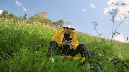 A young man in a yellow raincoat is sitting on a slope and texting on the phone in a mountainous area. Tourism and travel