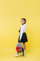 Smiling preteen pupil with backpack looking at camera on yellow background