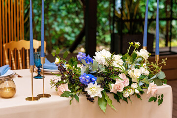 Wedding presidium, banquet table for newlyweds with flowers, greenery, blue and gold color. Lush flower arrangement on the wedding table. Glowing light bulbs. Soft selective focus.